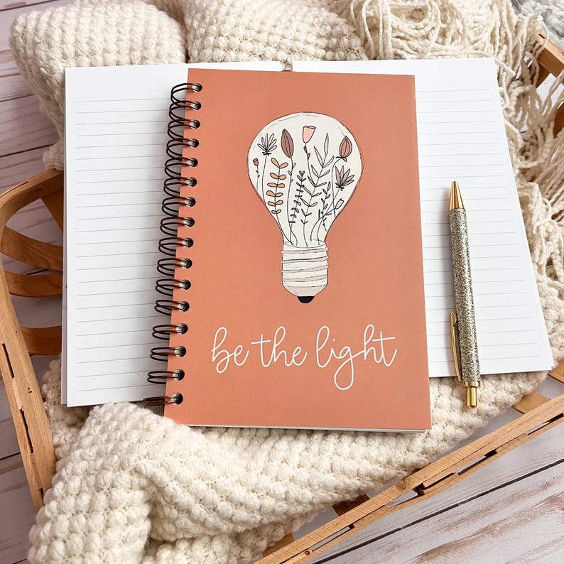 Puede incluir: Un cuaderno de espiral de color coral con una ilustraci&oacute;n de bombilla llena de flores y el texto "be the light".