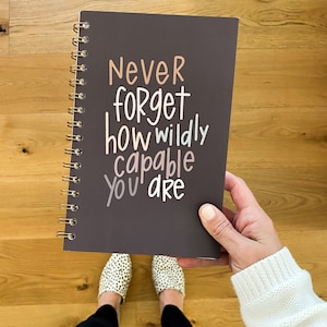 May include: A dark grey spiral bound journal with the text "Never forget how wildly capable you are" written in white with colourful accents.