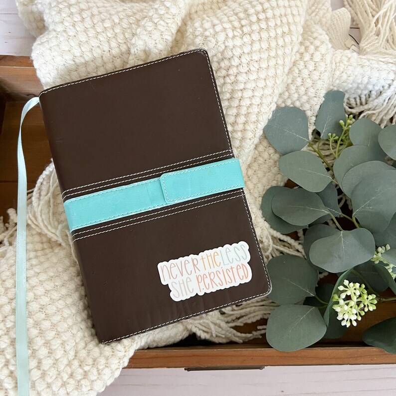 May include: A brown leather-bound journal with a turquoise ribbon bookmark. A sticker with the text "nevertheless she persisted" is on the cover.