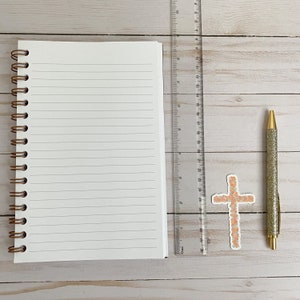 May include: A white spiral bound notebook with lined pages, a ruler, a pen with gold glitter, and a floral cross sticker on a light wood surface.
