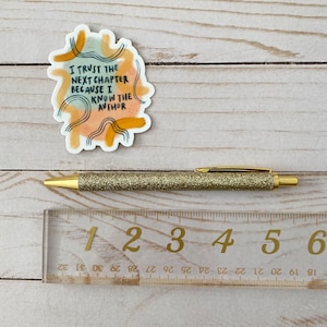 May include: A sticker with a colorful abstract design and the text "I trust the next chapter because I know the author". The sticker is on a wooden surface with a gold glitter pen and a ruler.