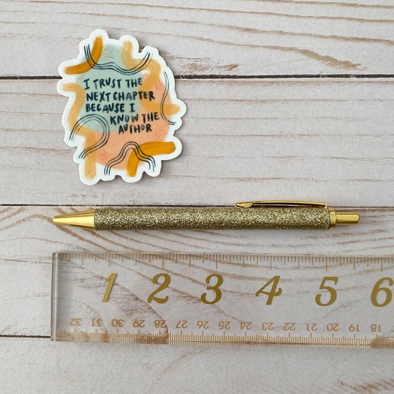 May include: A sticker with a colorful abstract design and the text "I trust the next chapter because I know the author". The sticker is on a wooden surface with a gold glitter pen and a ruler.