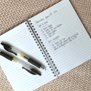 May include: A white notebook with black lines and a black spiral binding. The notebook has handwritten text on it, including the date "Thursday - June 3rd, 2021" and a list of tasks and activities. The text includes "get it done", "Bible Study", "yoga class", "photo all new stickers", "send orders", "call UPS", "girls schedule", "liv track til 5pm", "lily softball game 430 @ lee-field", "grace bball practice EBC @ 145".