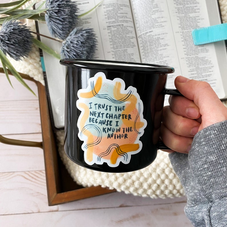 May include: Black enamel mug with a white sticker that says "I trust the next chapter because I know the author". The sticker has a colorful abstract design with yellow, orange, and blue accents.