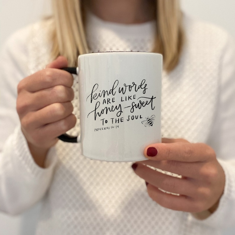 May include: White ceramic mug with a black handle. The mug has a black handwritten quote that reads "Kind words are like honey-sweet to the soul" with the scripture reference "Proverbs 16:24" below. A small black bee illustration is to the right of the quote.