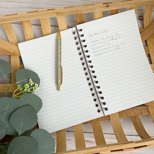May include: A white notebook with a brown spiral binding. The notebook is open to a page with a handwritten to-do list that includes "Bible study", "Grocery shop", "Dishes", "Dry cleaning", and "Exercise". A gold pen with glitter is lying on the page.