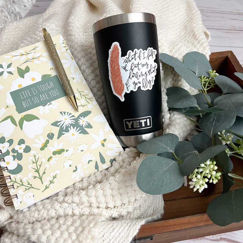 Op de afbeelding: Een zwarte Yeti-beker met een sticker waarop staat "what if I fall, oh but my darling what if you fly?" met een bruin verenontwerp. De beker staat op een houten oppervlak met een wit deken en een notitieboek met een bloemenpatroon.