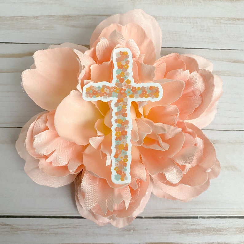 May include: A white cross sticker with a floral design in shades of pink, orange, and green. The sticker is placed on a pink flower.