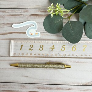 May include: A gold glitter pen with a clear ruler marked with numbers 1 through 7. The ruler is on a light wood surface with a blue and white sticker that says "Live Aloha".
