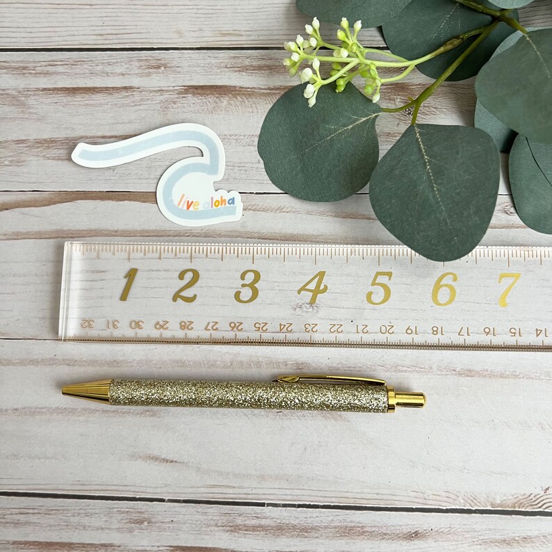 May include: A gold glitter pen with a clear ruler marked with numbers 1 through 7. The ruler is on a light wood surface with a blue and white sticker that says "Live Aloha".