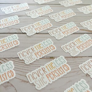 May include: A close-up of several stickers with the text "Never the less she persisted" in different colours and fonts. The stickers are on a light wood surface.