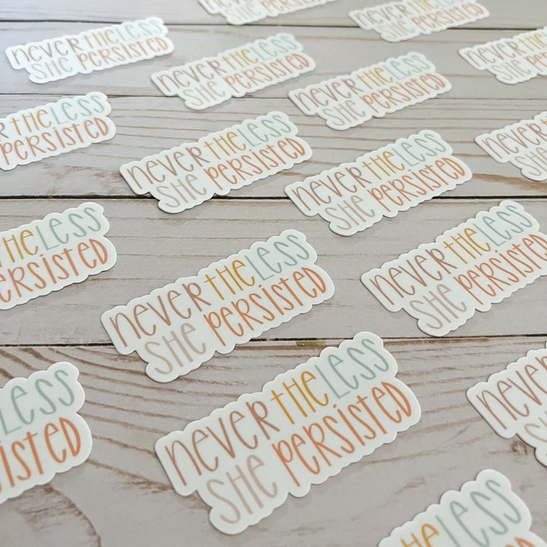 May include: A close-up of several stickers with the text "Never the less she persisted" in different colours and fonts. The stickers are on a light wood surface.