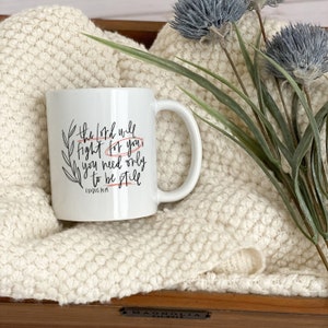 Op de afbeelding: Witte keramische mok met zwarte kalligrafie die "De Heer zal voor je vechten, je hoeft alleen maar stil te zijn" en "Exodus 14:14" zegt.