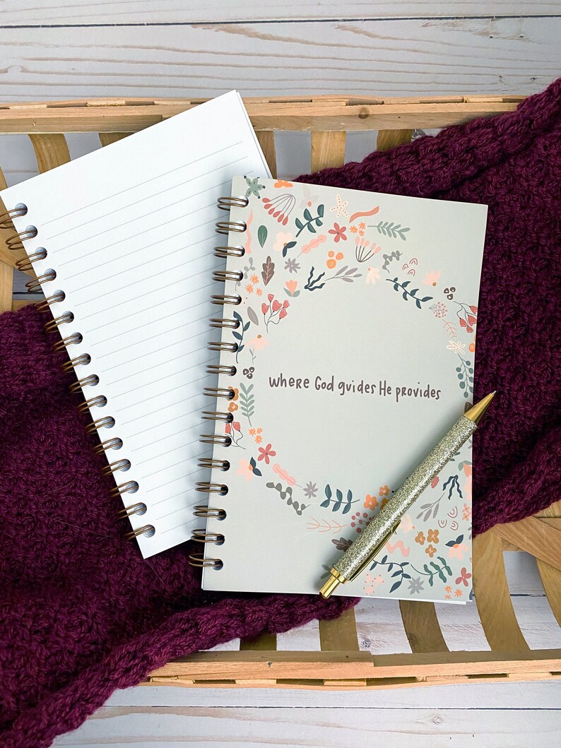 May include: A gray spiral bound journal with a floral design and the text "where God guides He provides". A gold pen is lying on top of the journal.