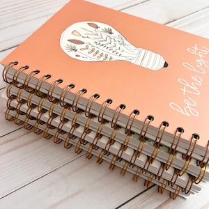 Puede incluir: Una pila de cuadernos de espiral con una cubierta de color melocot&oacute;n. La cubierta presenta una bombilla con dise&ntilde;os florales en su interior y el texto "S&eacute; la luz".