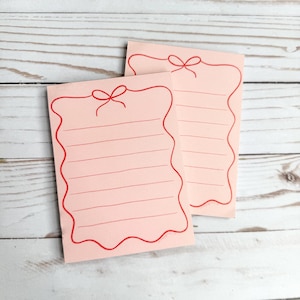 May include: Two pink notepads with red lines and a red bow design. The notepads have a wavy edge.