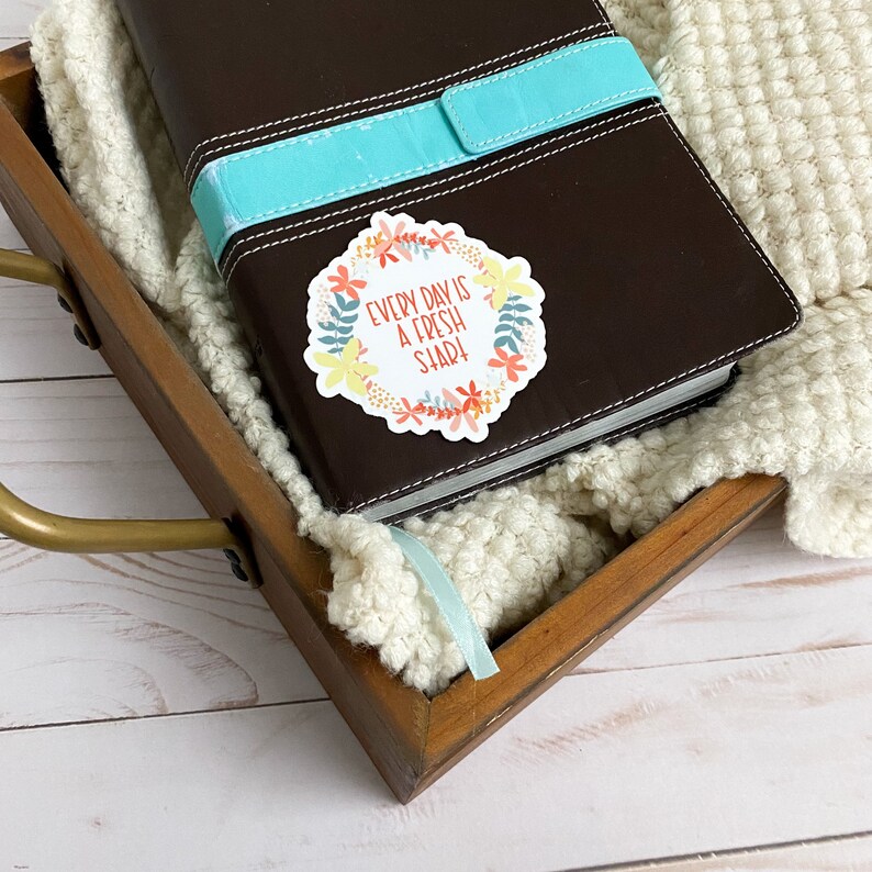 Peut inclure: Un journal en cuir marron avec une fermeture en ruban turquoise. Un autocollant floral avec le texte "Every day is a fresh start!" est sur la couverture.