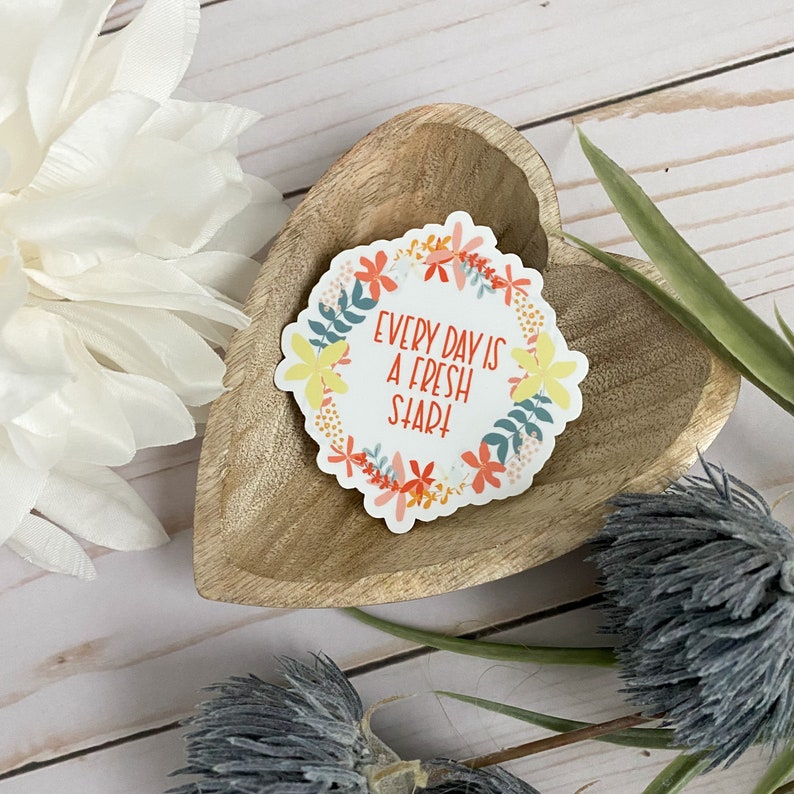 Peut inclure: Un autocollant avec un motif de couronne florale et le texte "Every day is a fresh start!" dans un cercle blanc. L'autocollant est sur un plat en bois en forme de c&oelig;ur.