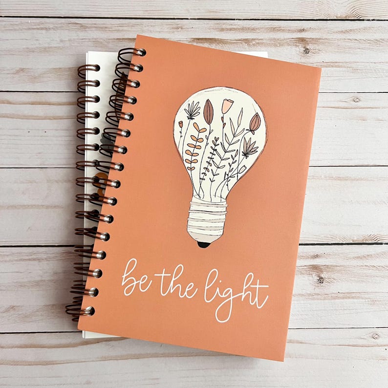 Puede incluir: Una libreta de espiral de color melocot&oacute;n con el texto "be the light" en escritura blanca. La portada presenta una bombilla con un dise&ntilde;o floral en su interior.