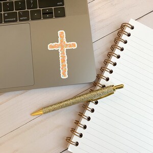 May include: A floral cross sticker on a silver laptop next to a spiral bound notebook and a gold glitter pen.