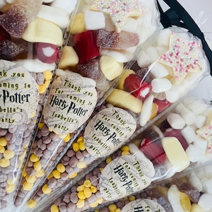 May include: A collection of clear plastic cones filled with various sweets, including yellow and brown balls, red and white sweets, and white marshmallows. Each cone has a round sticker with the text "Thank you for coming to Isabella's Harry Potter party".