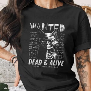 Puede incluir: Camiseta negra con un gráfico de un gato, mitad calavera, con gafas y el texto "WANTED DEAD & ALIVE SCHRODINGER'S CAT". El diseño incluye ecuaciones matemáticas. La camiseta es de color oscuro.