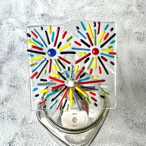 May include: A square glass night light featuring a colorful fused glass design of three firework bursts. The fireworks are composed of thin, raised lines in red, yellow, blue, green, black, and white, radiating from central circular points.