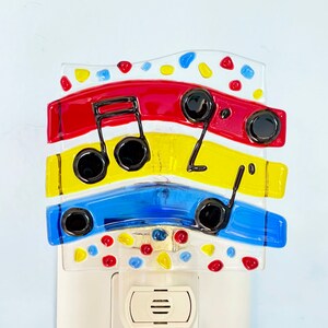May include: A colorful night light featuring a musical note design. The night light has layers of red, yellow, and blue glass with black musical notes. The top layer is white with red, yellow, and blue accents. The night light plugs into a standard wall outlet.