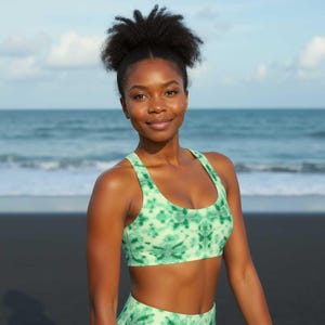 May include: A woman wearing a green and white tie-dye sports bra and matching leggings. She is standing on a black sand beach with the ocean in the background.