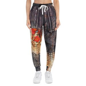 May include: Jogger sweatpants with a distressed, textured design in shades of brown, black, and orange. The left leg features a skull and lightning bolt graphic. The pants have a drawstring waist and ribbed cuffs.