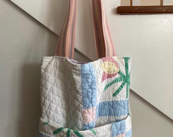 May include: A quilted tote bag with a floral design in pink, blue, and green. The bag has a pocket on the front and striped pink and yellow straps. The bag is hanging on a white wall.