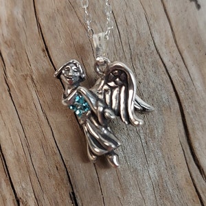 May include: A silver angel pendant necklace with a blue gemstone in the center. The angel is holding the gemstone in its hands and has wings spread out behind it.