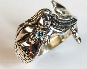 Puede incluir: Un anillo de plata con un diseño de sirena detallado y una piedra preciosa azul. La sirena sostiene la piedra preciosa en sus manos y su cola está enroscada alrededor de la banda del anillo.