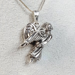 May include: Silver pendant necklace featuring a detailed angel with wings outstretched. The angel is depicted in a flying pose, with its head tilted downwards.