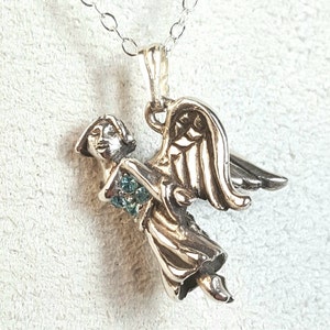 May include: Silver angel pendant with a detailed design and a small cluster of light blue gemstones. The angel is wearing a flowing gown and has wings outstretched.