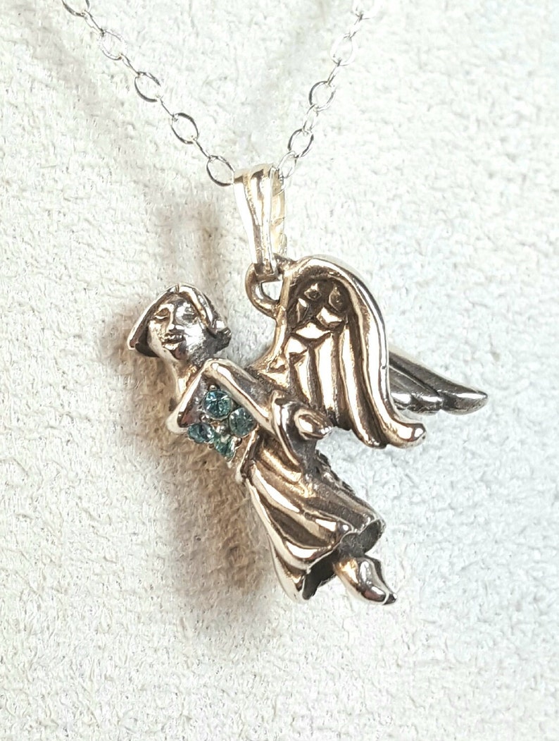 May include: Silver angel pendant with a detailed design and a small cluster of light blue gemstones. The angel is wearing a flowing gown and has wings outstretched.