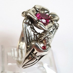 May include: A silver ring featuring a floral design with a large, oval, ruby-colored gemstone at the center. The ring has an intricate design with a figure and smaller red gemstone accents.