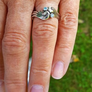 May include: A silver ring with a swirling design and three small, round, light blue gemstones. The ring is worn on a finger, with the hand and a blurred green background visible.
