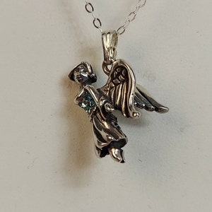 May include: Silver angel pendant with a green gemstone on a silver chain. The angel is depicted with wings and a flowing robe.