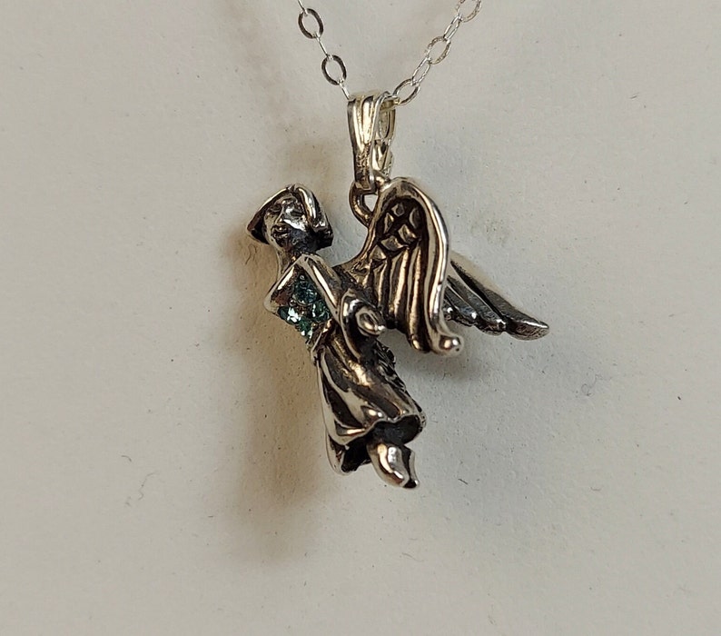 May include: Silver angel pendant with a green gemstone on a silver chain. The angel is depicted with wings and a flowing robe.
