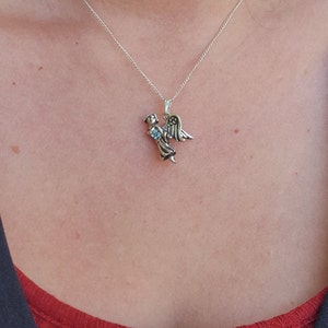 May include: A silver necklace with a small angel pendant. The angel is holding a blue gemstone in its hands.