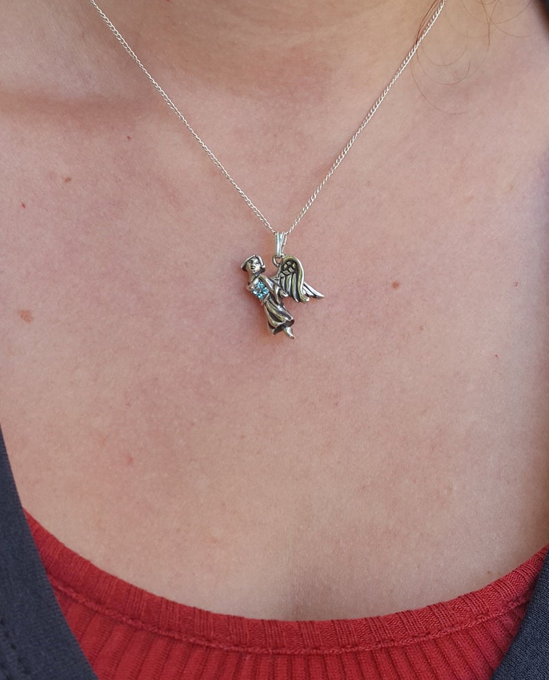 May include: A silver necklace with a small angel pendant. The angel is holding a blue gemstone in its hands.