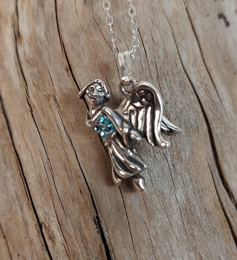 May include: A silver angel pendant necklace with a blue gemstone in the center. The angel is holding the gemstone in its hands and has wings spread out behind it.