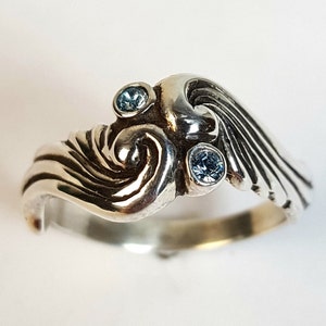 May include: A sterling silver ring with a wave design, featuring three small, round, light blue gemstones. The ring has a swirling, textured pattern and a polished finish, suitable for everyday wear.