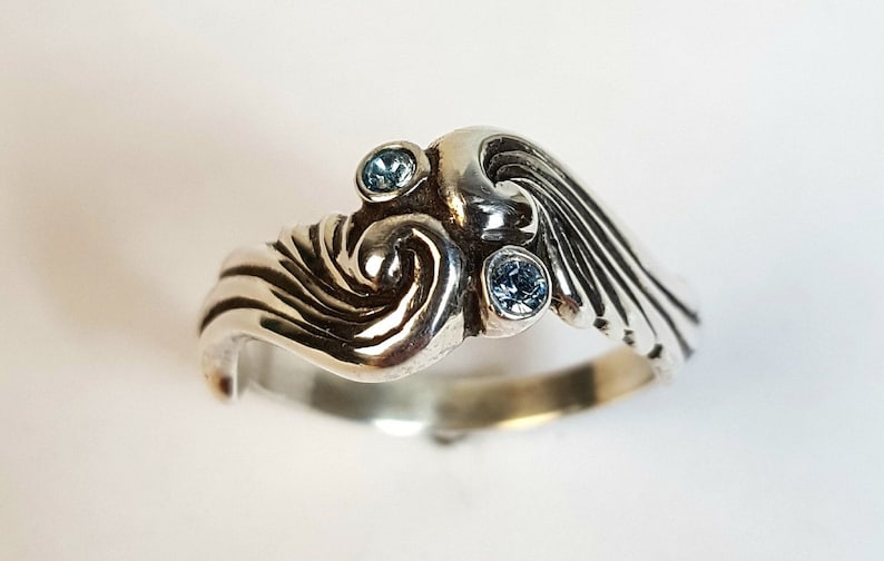 May include: A sterling silver ring with a wave design, featuring three small, round, light blue gemstones. The ring has a swirling, textured pattern and a polished finish, suitable for everyday wear.