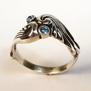 May include: A silver ring with an Art Nouveau design, featuring a stylized face and flowing lines. The ring is adorned with three small, round, light blue gemstones. The ring is set against a white background.