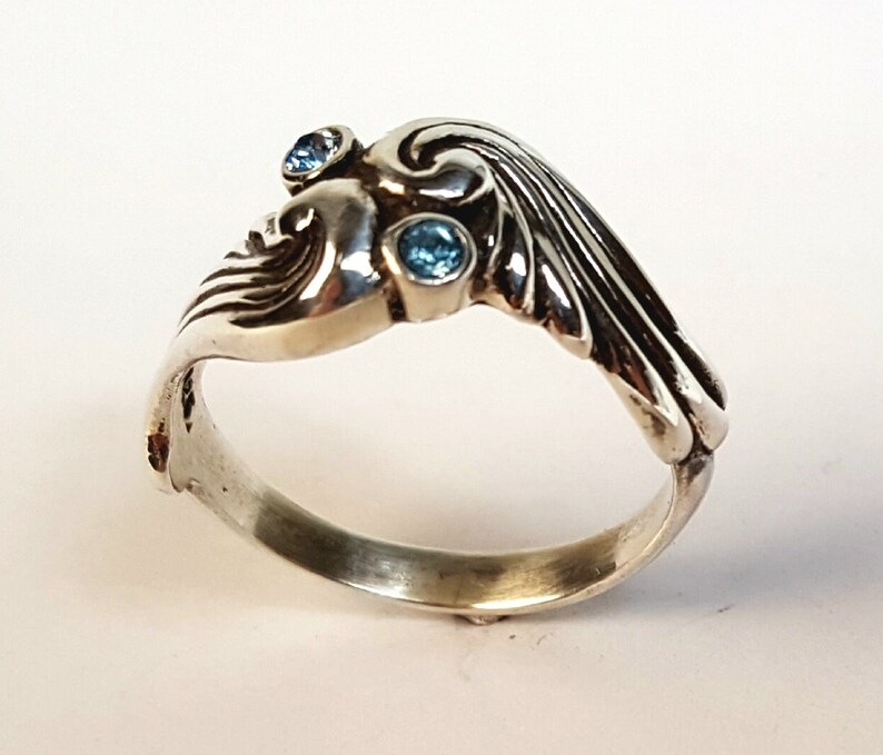 May include: A silver ring with an Art Nouveau design, featuring a stylized face and flowing lines. The ring is adorned with three small, round, light blue gemstones. The ring is set against a white background.