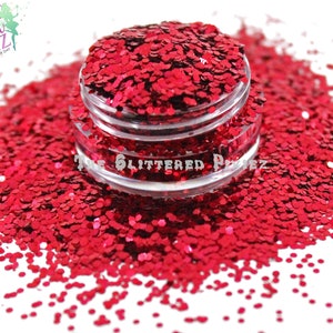 May include: A jar of vibrant red glitter, spilling onto a white surface. The glitter is in small, irregular shapes, creating a textured appearance. The jar has a clear plastic body and a silver lid. The text "The Glittered Pixiez" is visible.
