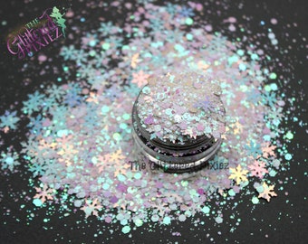 May include: A close-up of a jar of iridescent glitter with a mix of snowflake and circle shapes in shades of white, blue, and purple. The glitter is scattered around the jar, creating a sparkling effect.