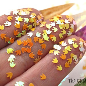 May include: A close-up of iridescent orange and yellow flower-shaped glitter. The glitter is scattered on a white surface and is being held in a hand.